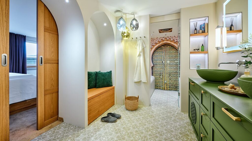 Von Hammams inspiriert: Warmgrünes Sideboard mit „Blume des Lebens“, runde Aufsatzbecken, Betonfliesen mit Ornamenten in Naturtönen, dazu die in Beige gehaltenen Wände und von typischen Spitzbögen geprägte Nischen; eingepasst sind Regale und Auszüge aus Eiche, einer davon als Sitzbank. Eine Schiebetür verbindet den Bereich mit dem Schlafzimmer. Bild: MHK Group