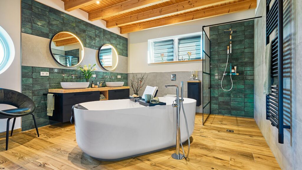 Spiegel nehmen die Form des Rundfensters auf; Bodenfliesen in Eichenoptik greifen den Holzton von Decke und Waschtischabdeckung auf; tiefes Petrol an den Rückwänden von Dusche und Waschplatz setzt Akzente, aufgehellt durch sandfarbene Fliesen. Schwarze Details wie Duschabtrennung, Handtuchwärmer und Möbel verleihen industriellen Charakter. Bild: MHK Group