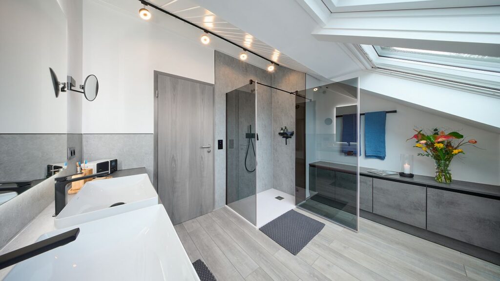 Dachschräge durch Maßanfertigungen gut genutzt: Ein wandbündiger Waschtisch mit zwei Aufsatzbecken bietet Ablagefläche, ein dimmbarer Lichtspiegel fügt sich in die Schräge ein. Gegenüber dient ein flaches Sideboard als Sitzplatz und Wäschesammler, inklusive versteckter Revisionsöffnung. Bild: MHK Group